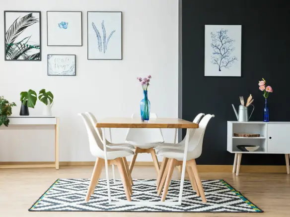 A modern dining area in Ulcinj, Montenegro real estate properties, featuring a wooden table, four white chairs, and a blue vase with flowers. A black-and-white rug covers the floor beneath framed botanical prints and minimal decorations by ekosphere.