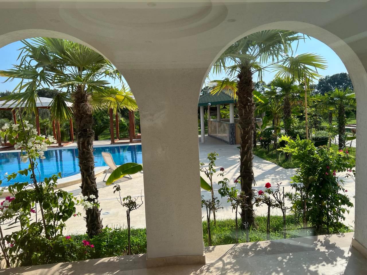 Stunning Ulcinj villa for sale: tropical garden, palm trees, pool view through white arches. Invest via immobilien.ekosphere.me by Ekosphere Doo.