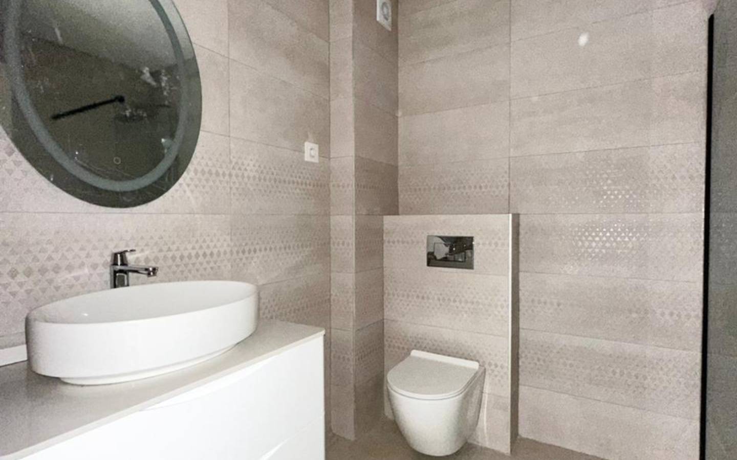 Stylish modern bathroom in luxury Ulcinj property: beige tiles, wall-mounted toilet, round mirror, and chic vanity. immobilien.ekosphere.me by ekosphere doo.