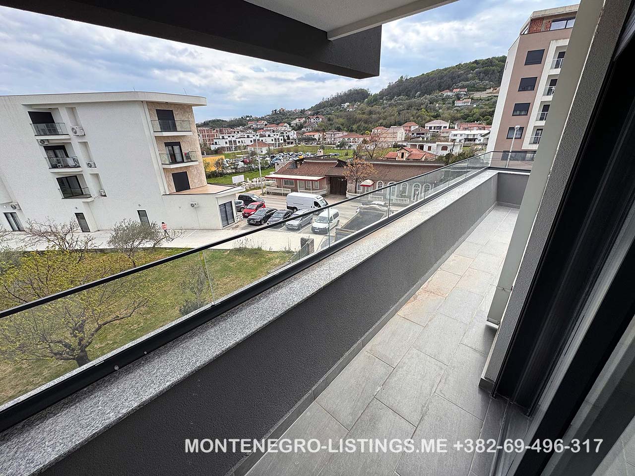 Stunning balcony view with glass railing over prime Ulcinj real estate&mdash;buildings, cars, lush hills. immobilien.ekosphere.me by Ekosphere doo.