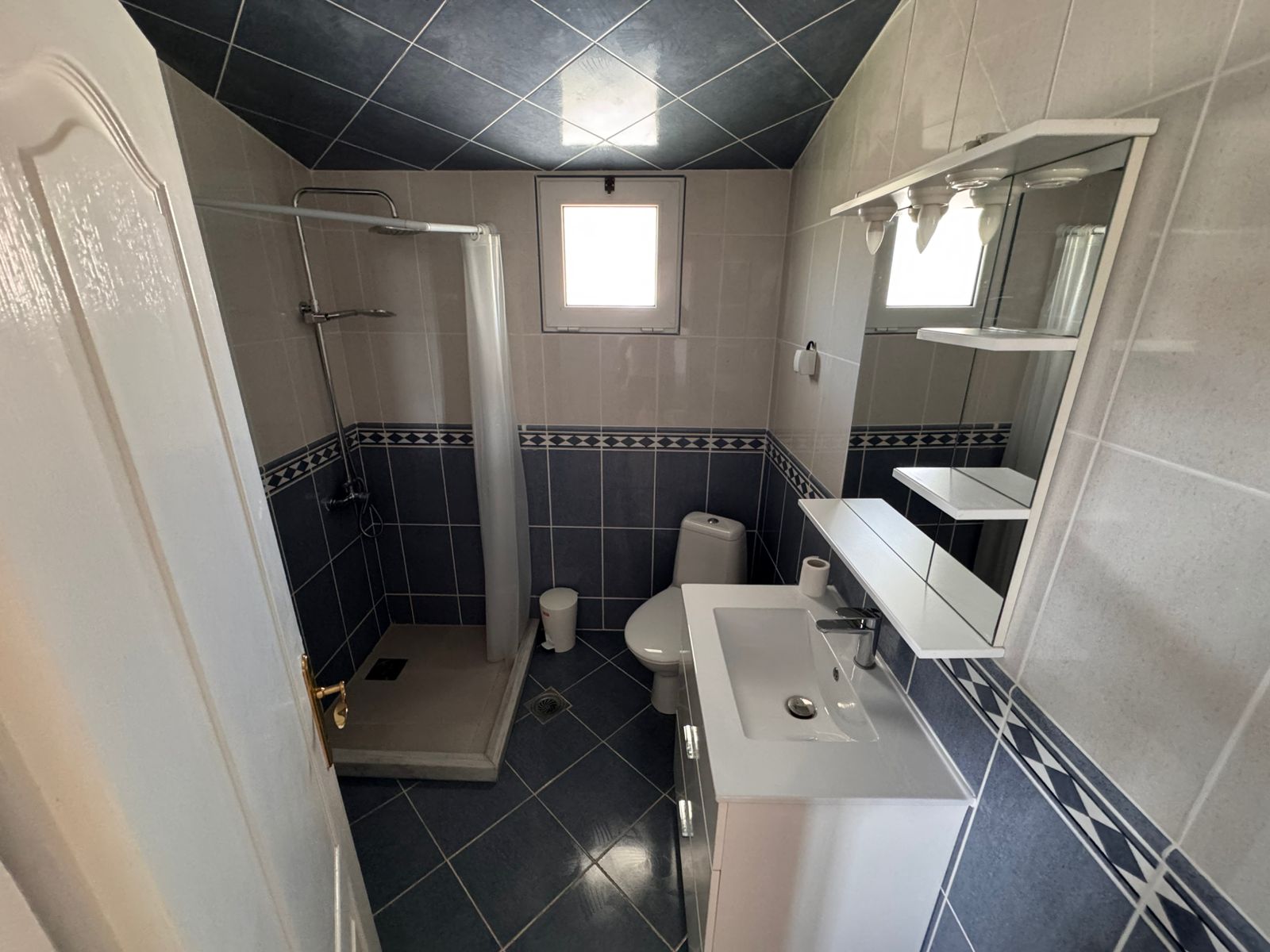 Modern Ulcinj Montenegro property bathroom: blue-white tiles, glass shower, window. Invest with immobilien.ekosphere.me by ekosphere doo.