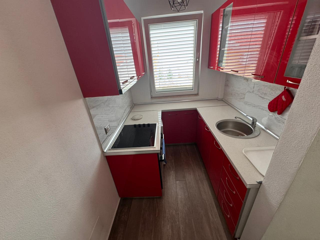 Modern Ulcinj kitchen: red cabinets, white countertop, electric stovetop. Ideal investment. Listed by immobilien.ekosphere.me, Ekosphere Doo.