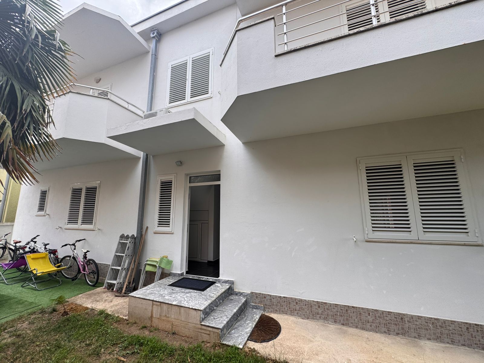 Modern white two-story home in Ulcinj, with porch, open entry, child-friendly lawn by palm tree. Listed on immobilien.ekosphere.me by ekosphere doo.