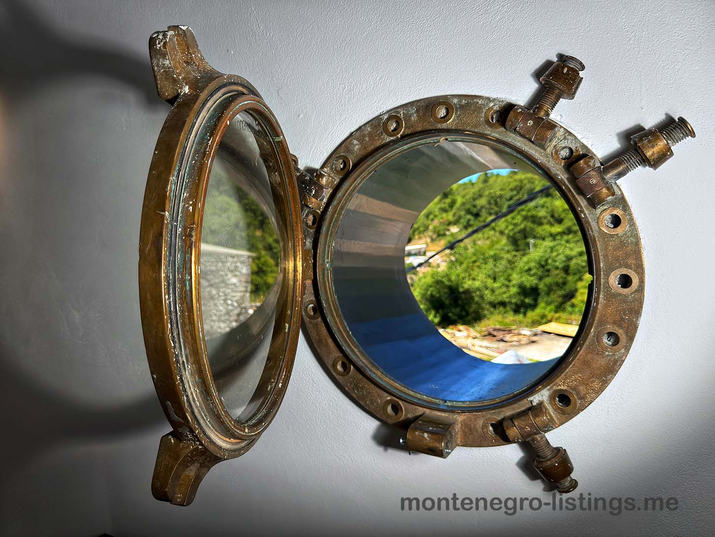 Open brass porthole on white wall reveals lush Ulcinj views&mdash;explore top Montenegro real estate at immobilien.ekosphere.me by ekosphere doo.