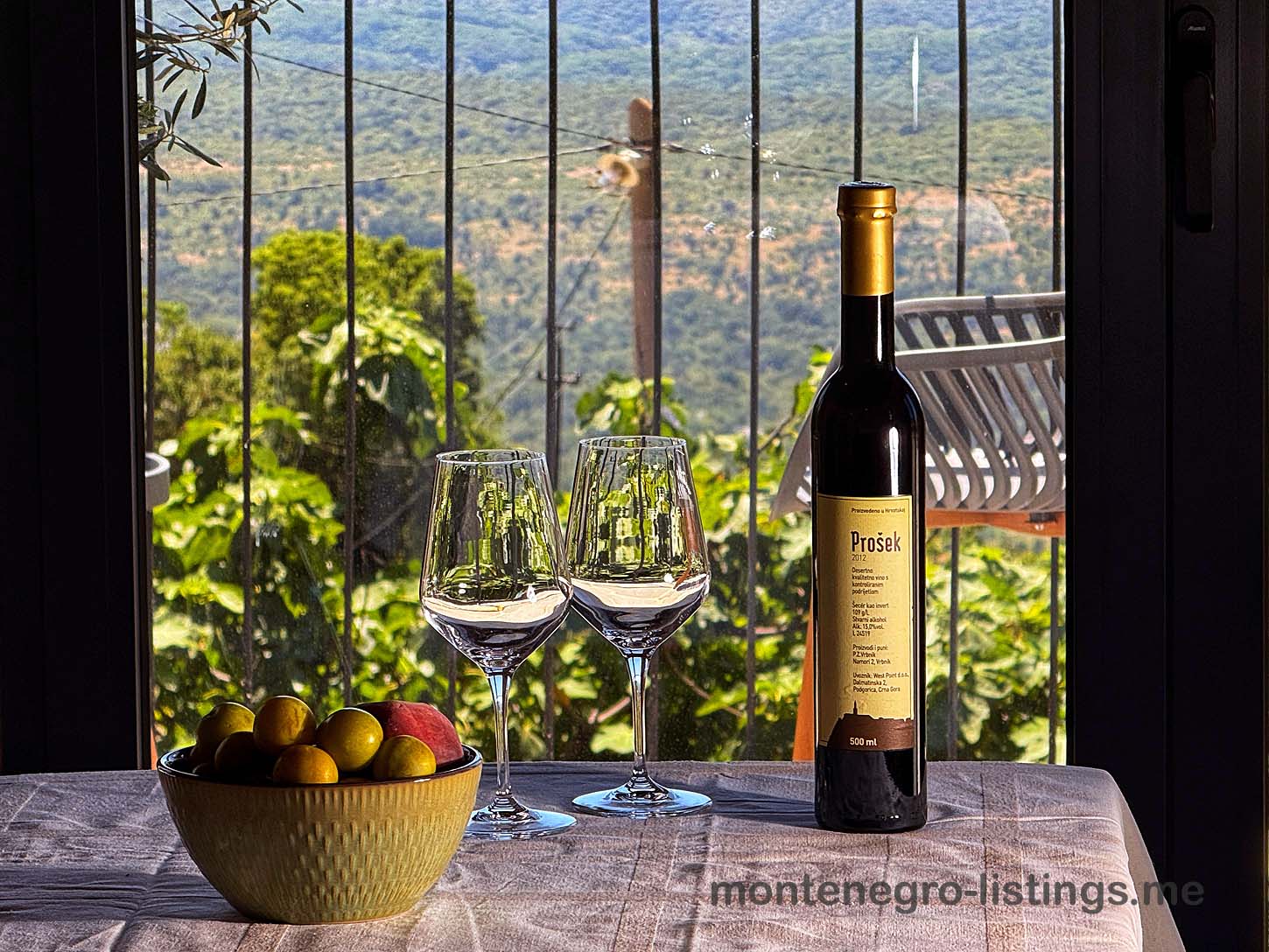 Pro&scaron;ek wine, glasses, and fresh fruit set by a window with green hill views&mdash;experience Ulcinj real estate luxury with immobilien.ekosphere.me.