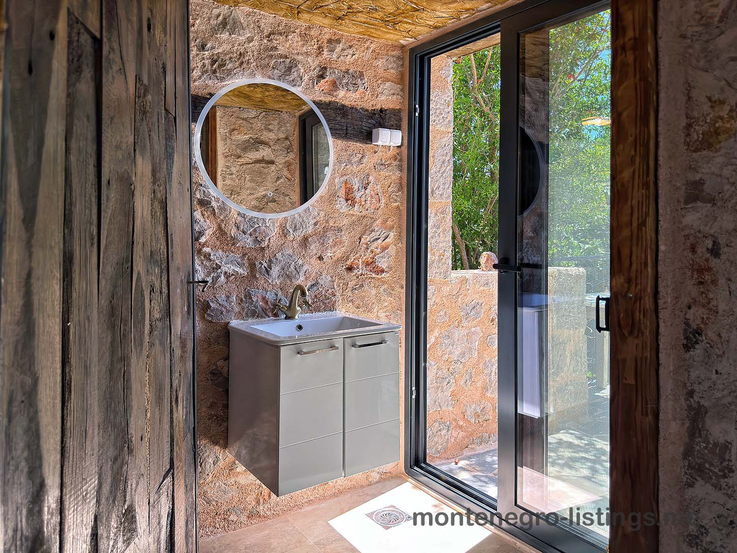 Luxury modern bathroom in Ulcinj, Montenegro: elegant gray vanity, stone walls, garden view. Invest with immobilien.ekosphere.me by Ekosphere.