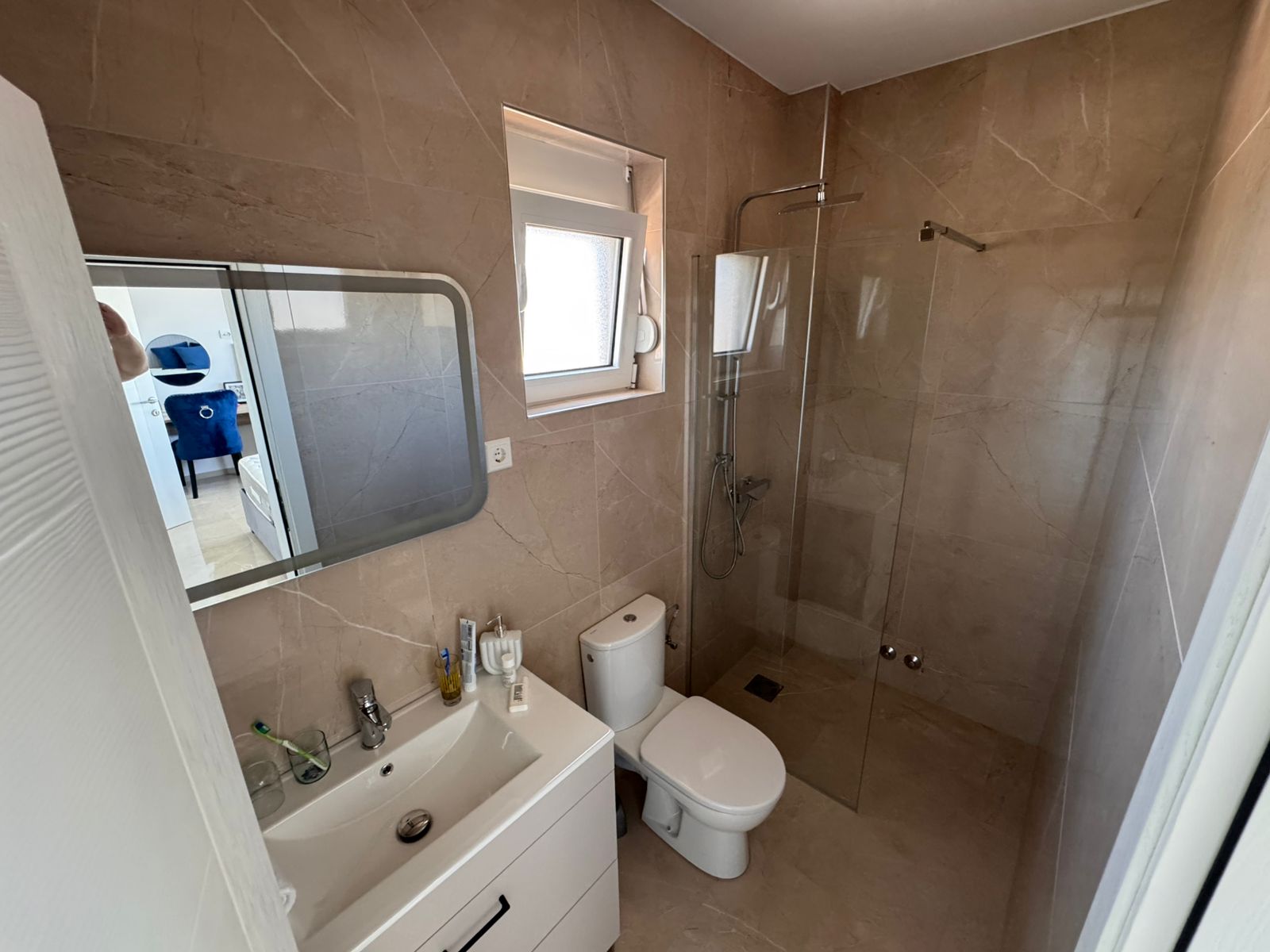 Elegant Ulcinj bathroom with beige tiles, glass shower, modern vanity; bright bedroom view. Invest with immobilien.ekosphere.me (ekosphere doo).