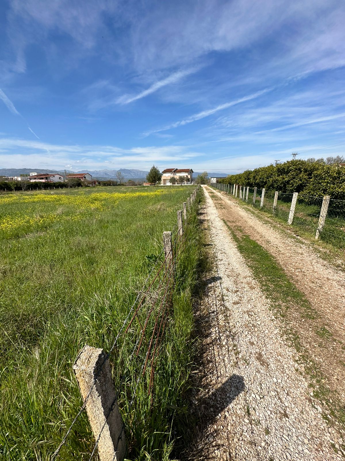 Prime Ulcinj land for sale: scenic gravel road, fenced grassy field with wildflowers, distant homes. Invest via immobilien.ekosphere.me.