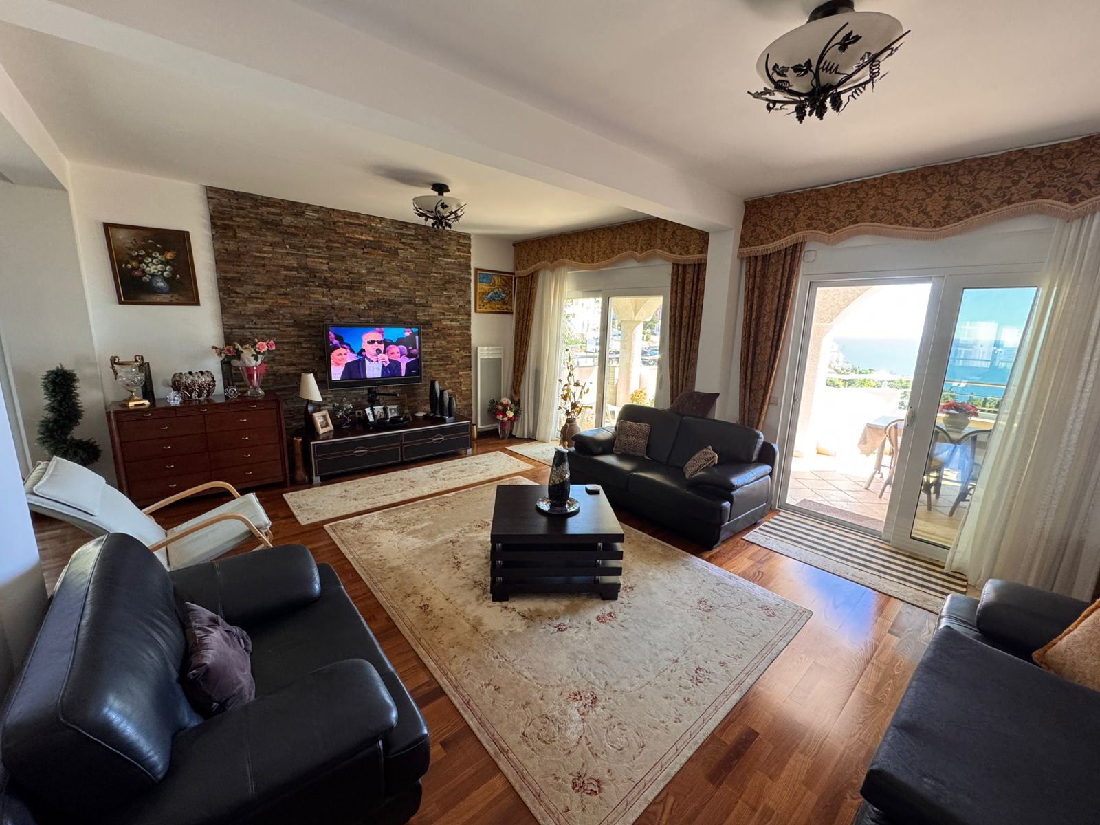 Elegant Ulcinj living room: black leather sofas, TV, stone wall, sea-view balcony. Invest via immobilien.ekosphere.me by ekosphere doo.