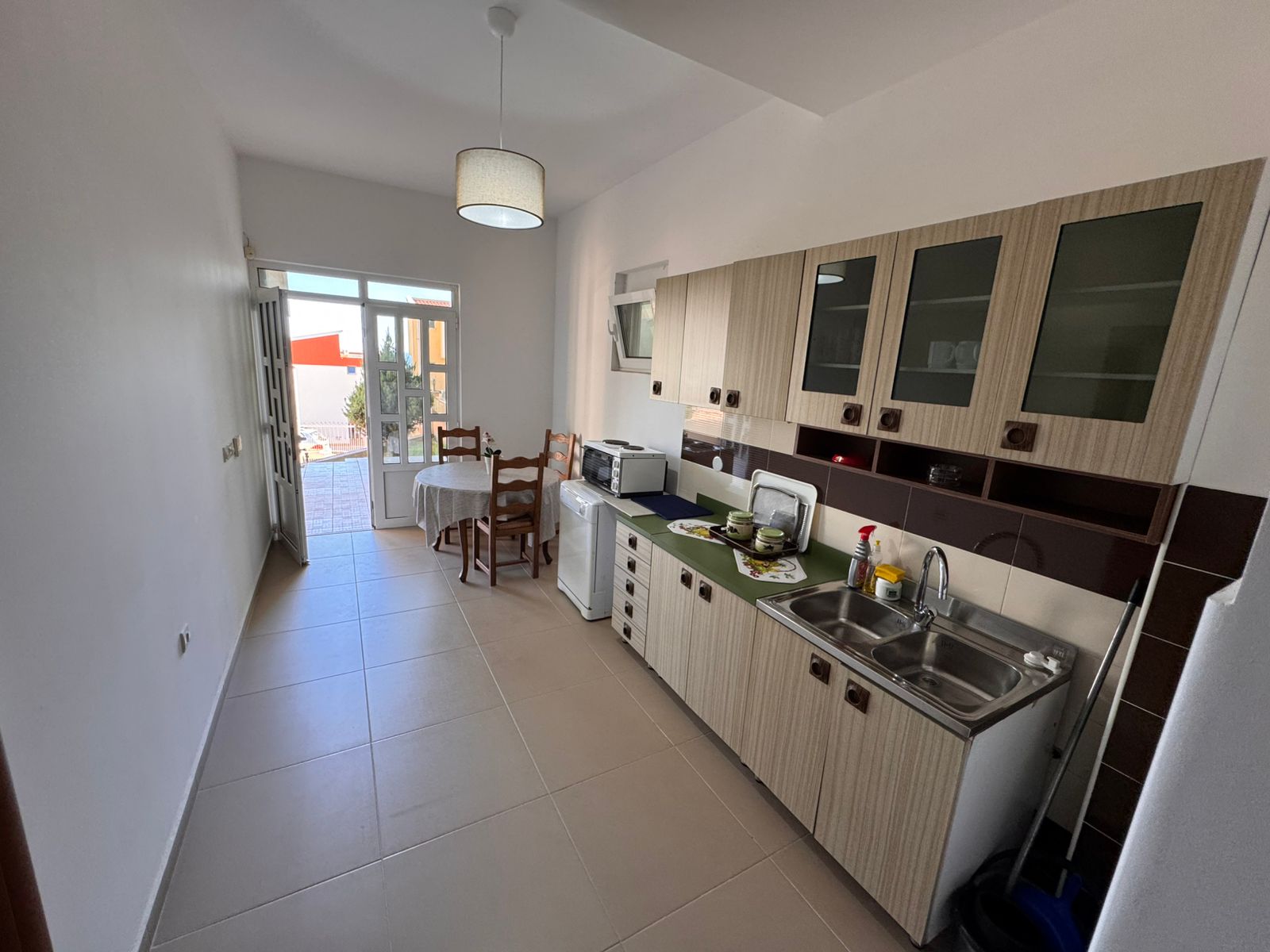 Modern Ulcinj kitchen with bright cabinets, double sink, dining area, and glass door to outdoor space&mdash;exclusive at immobilien.ekosphere.me.