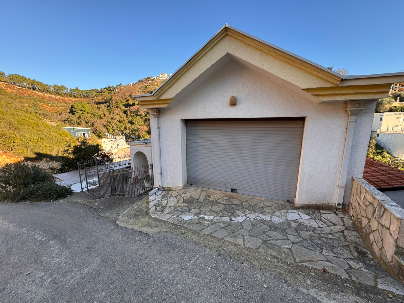 Modern white garage with secure gray door on a sloped lot, stone-paved drive, scenic Ulcinj views&mdash;exclusive at immobilien.ekosphere.me.