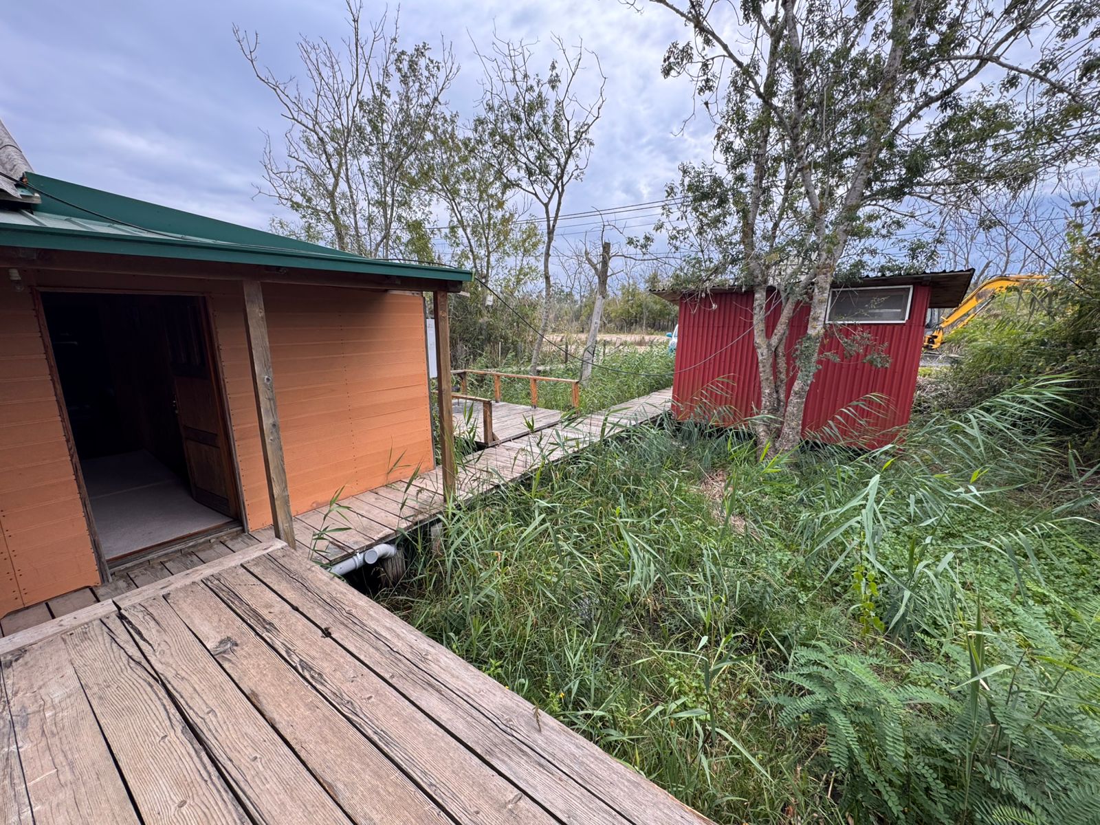 Charming Ulcinj property: wooden boardwalk links rustic brown home to red shed amid lush greenery. Invest via immobilien.ekosphere.me by ekosphere doo.