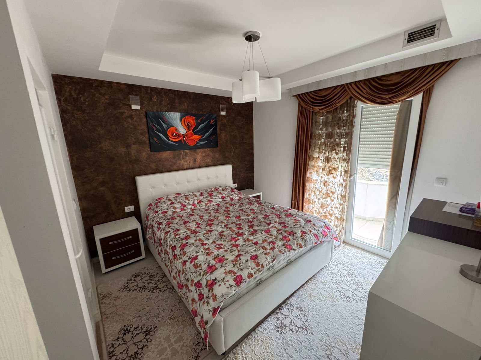 Modern Ulcinj bedroom with white bed, floral bedding, elegant nightstands, accent wall art, and large sunlit windows&mdash;exclusive at immobilien.ekosphere.me by ekosphere doo.