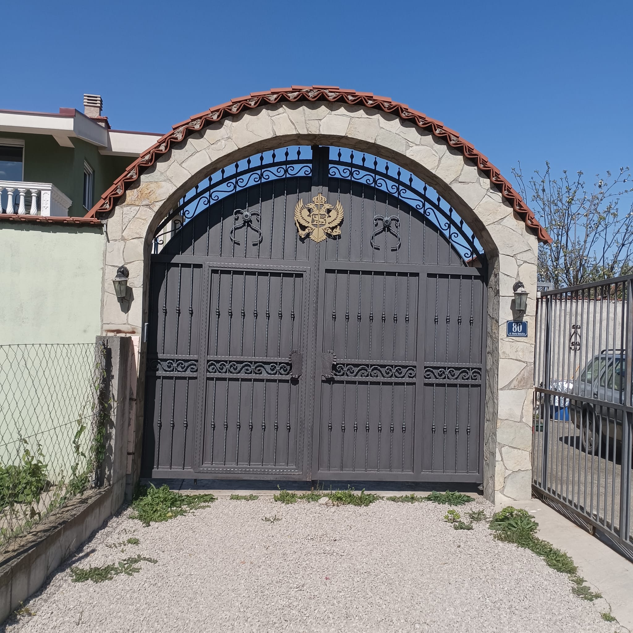 Elegant metal gate with golden crest, stone arch, dual lanterns, and gravel drive&mdash;exclusive Ulcinj real estate by ekosphere doo.