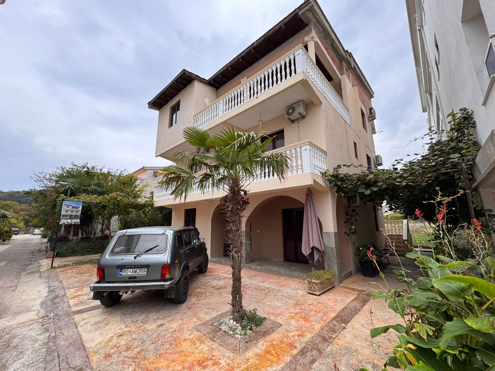 Elegant three-story beige villa with balconies, lush greenery, palm tree, and private driveway in Ulcinj. Listed on immobilien.ekosphere.me by ekosphere doo&mdash;prime real estate investment opportunity!.